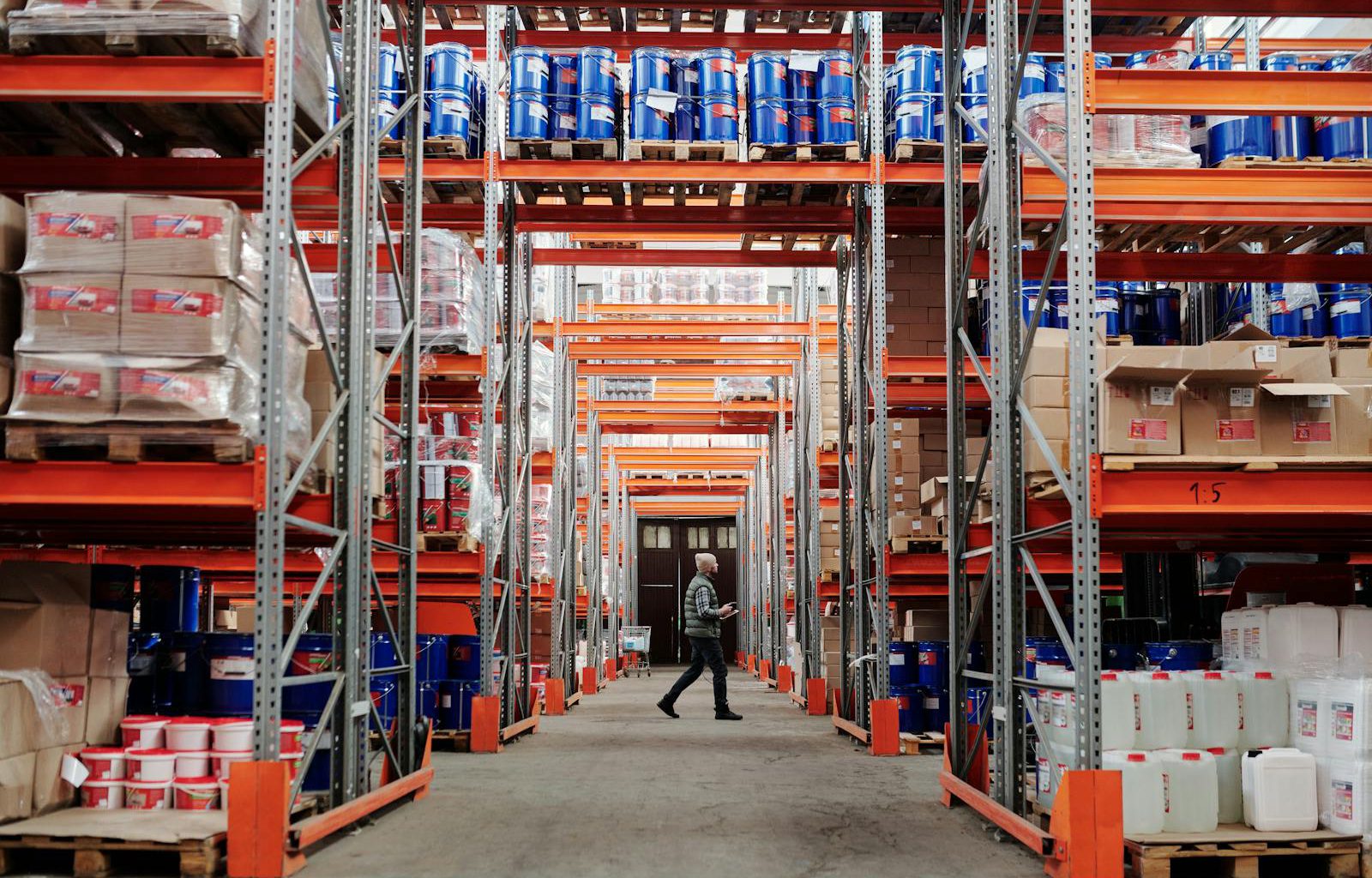 A man walking through a large industrial warehouse with stacked shelves filled with goods and products.