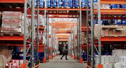 A man walking through a large industrial warehouse with stacked shelves filled with goods and products.