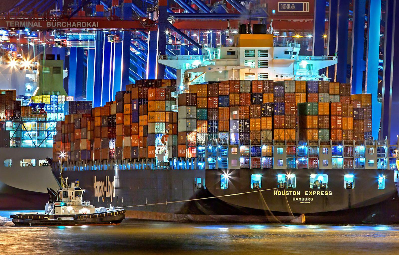 A brightly lit cargo ship at Hamburg harbor with stacked containers and a tugboat.