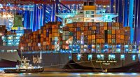 A brightly lit cargo ship at Hamburg harbor with stacked containers and a tugboat.