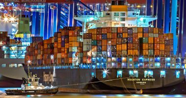A brightly lit cargo ship at Hamburg harbor with stacked containers and a tugboat.