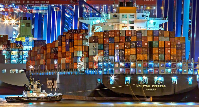 A brightly lit cargo ship at Hamburg harbor with stacked containers and a tugboat.